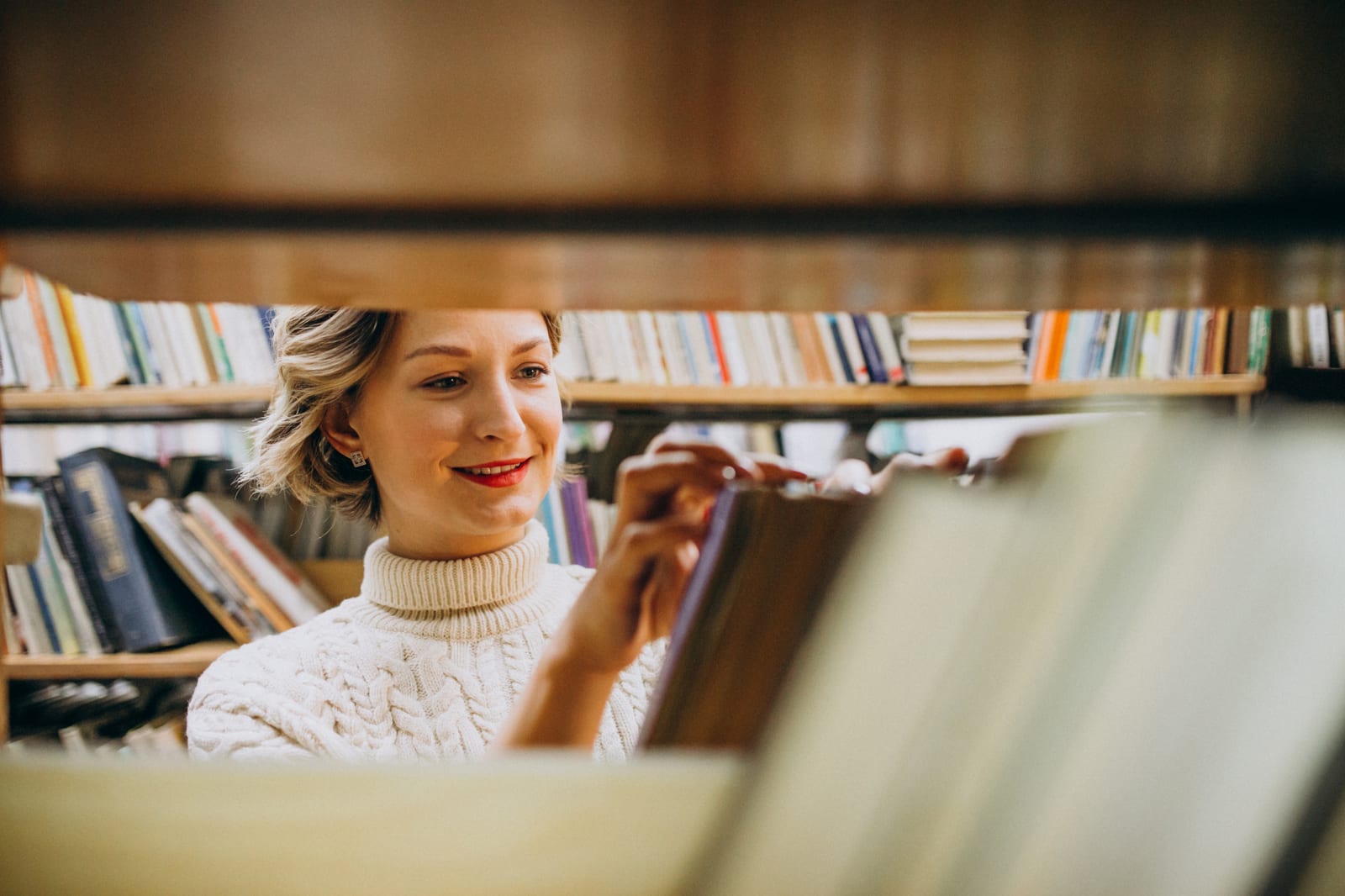 Mulher sorri ao retirar um livro de uma estante repleta de volumes, vista por entre as prateleiras.