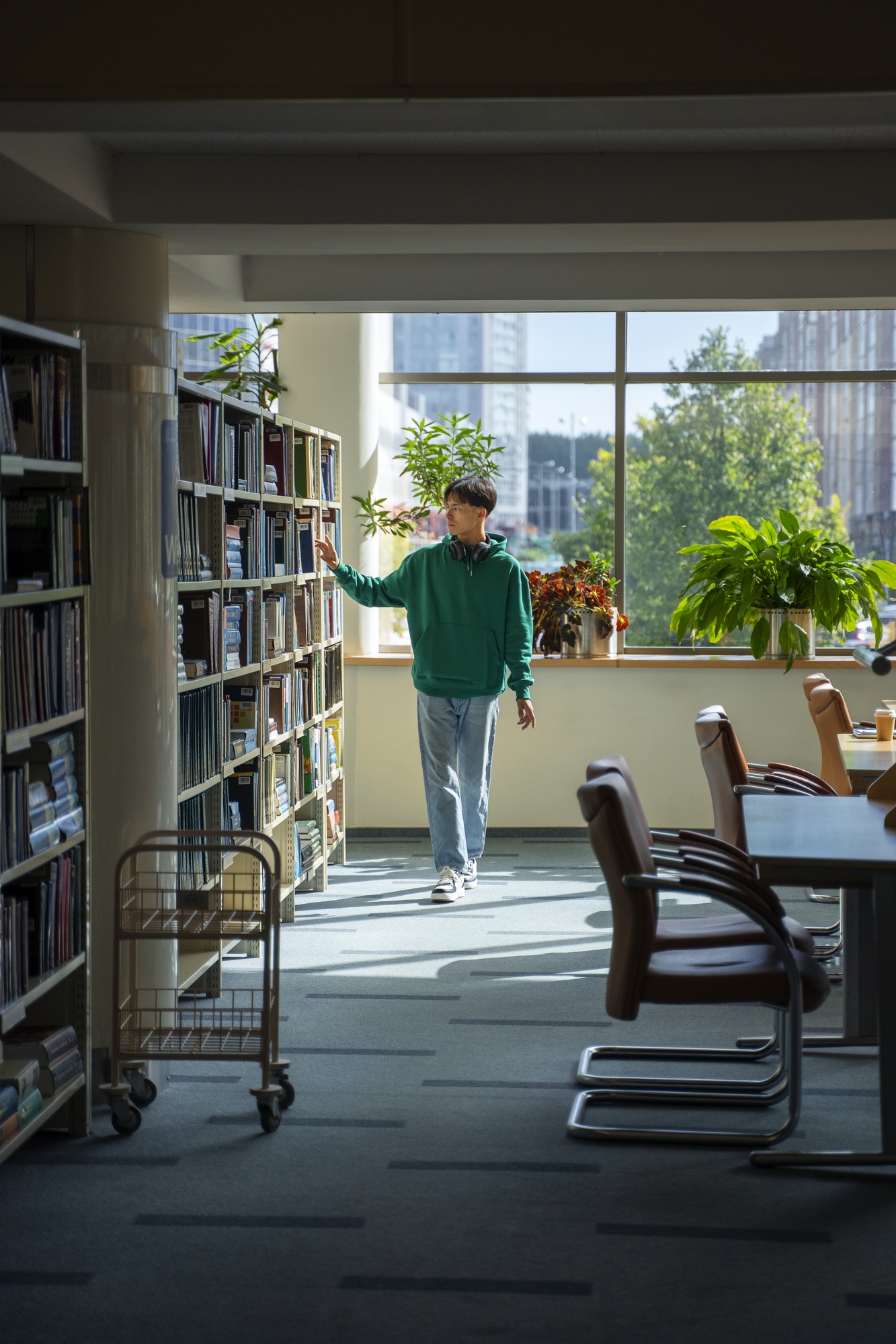Homem percorre um corredor de estantes em uma biblioteca, observando os livros organizados nas prateleiras.