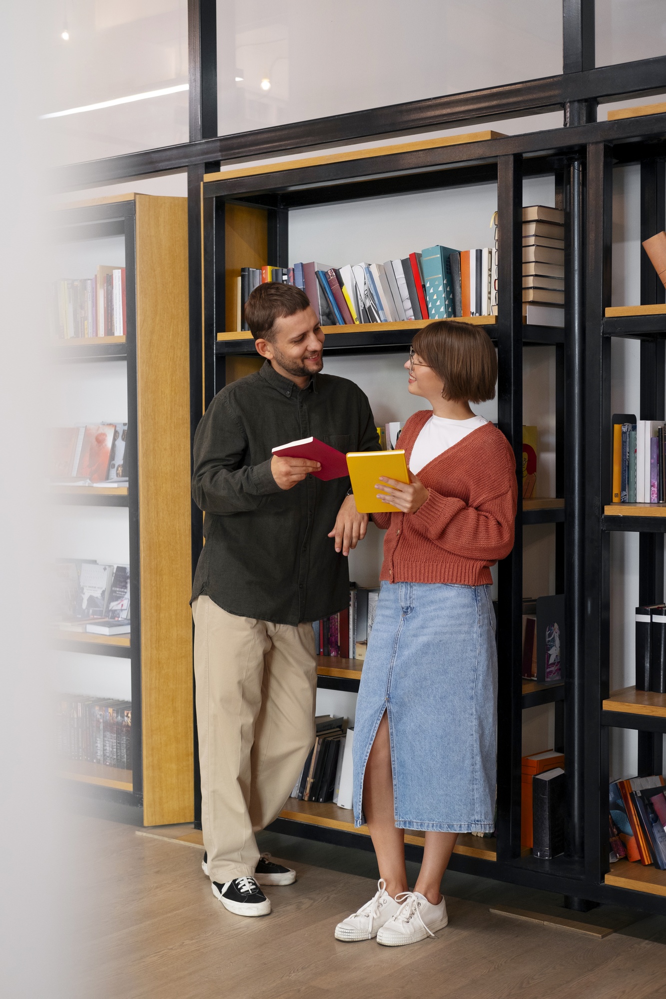 Duas pessoas sorriem enquanto trocam livros de capas coloridas diante de uma estante repleta de volumes.